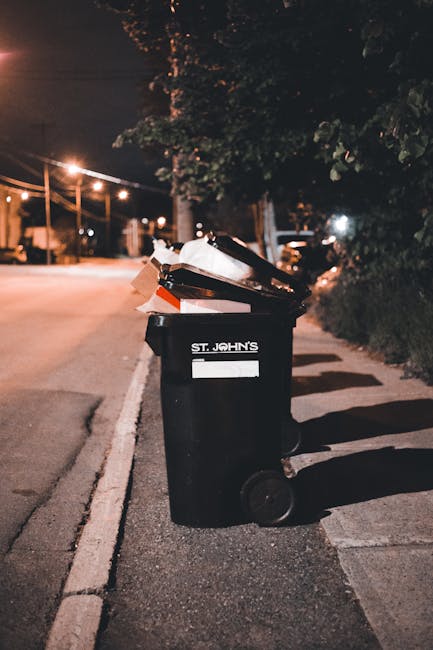 A pile of assorted waste materials placed on a paved driveway, including large cardboard boxes, black plastic bin bags, and wooden planks, all showing signs of wear and dirt. The cardboard boxes are s