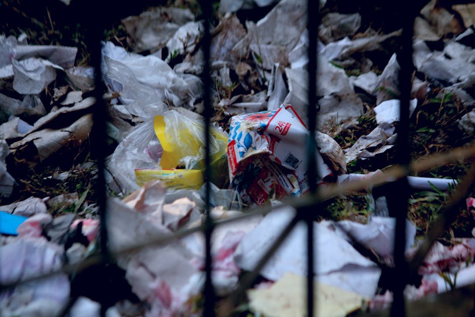 A close-up view of a pile of domestic waste visible through black metal fence bars. The rubbish consists mainly of crumpled and torn plastic bags, some white and yellow, with various paper and cardboa